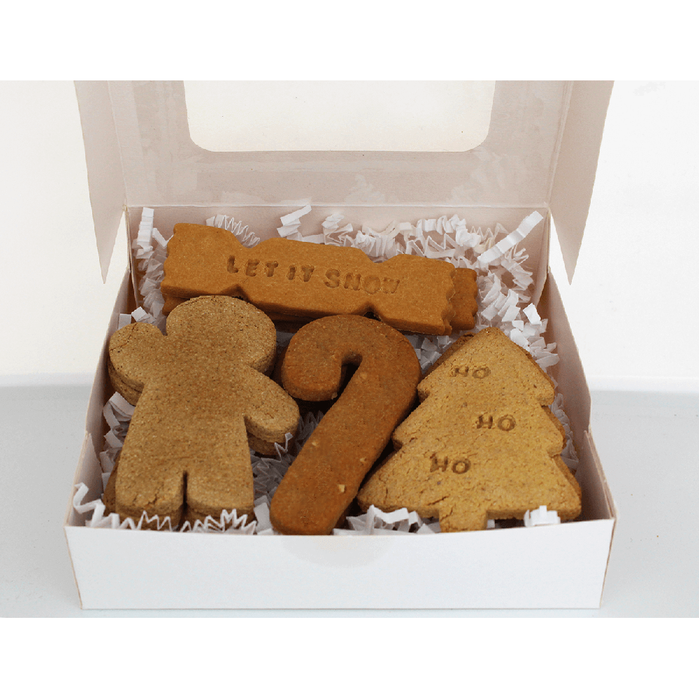 A white gift box with a clear window lid, filled with holiday cookies shaped like a gingerbread man, a candy cane, a Christmas tree, and a snowman, set against a plain white background.